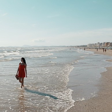 Una ragazza con un vestito rosso cammina a piedi nudi lungo il bagnoasciuga tenendo con la mano destra le scarpe.